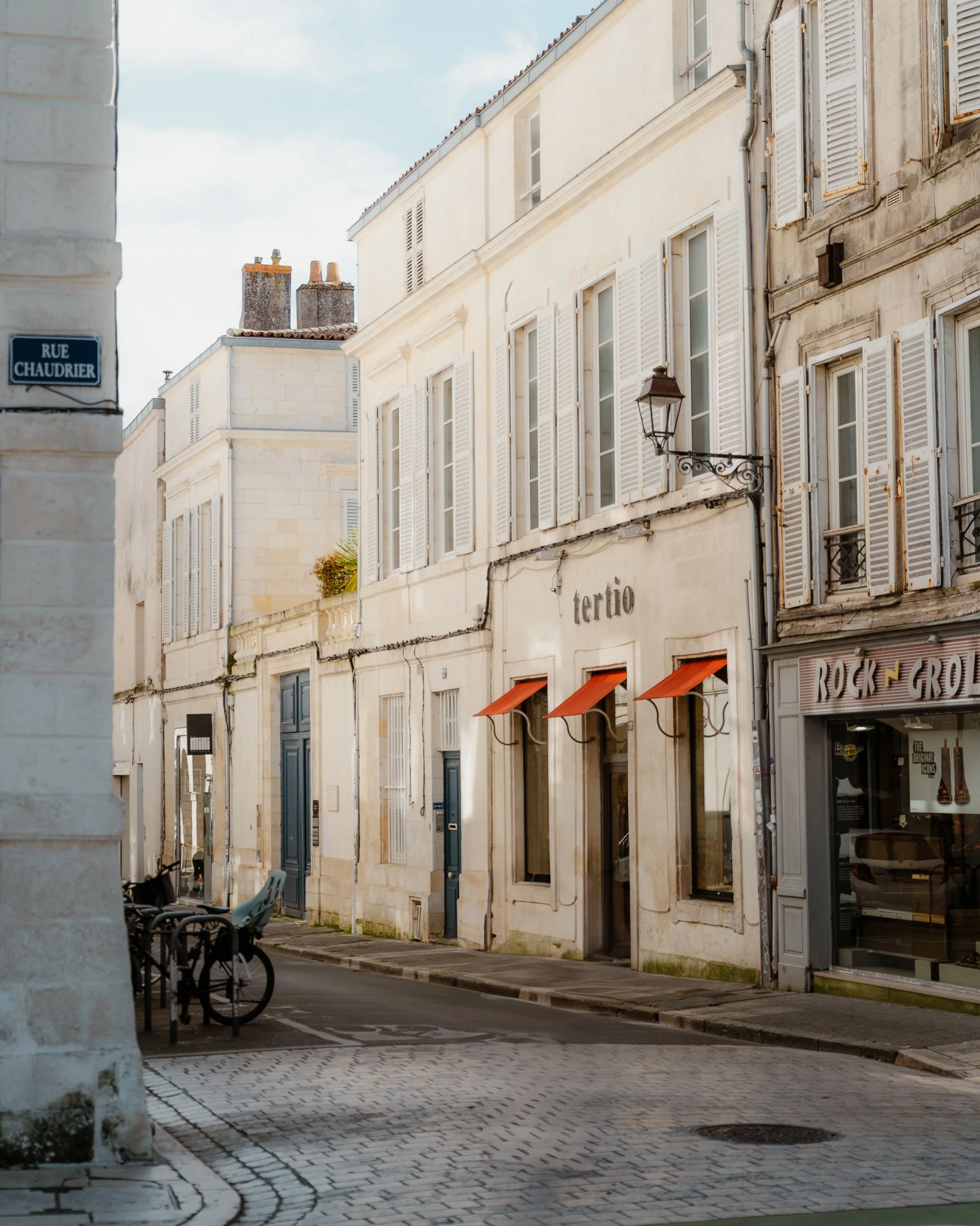 A quiet European street with white buildings, some with open window shutters, and a sign that reads 'tertio' above an entrance with orange awnings. There are bicycles parked on the cobblestone sidewalk.