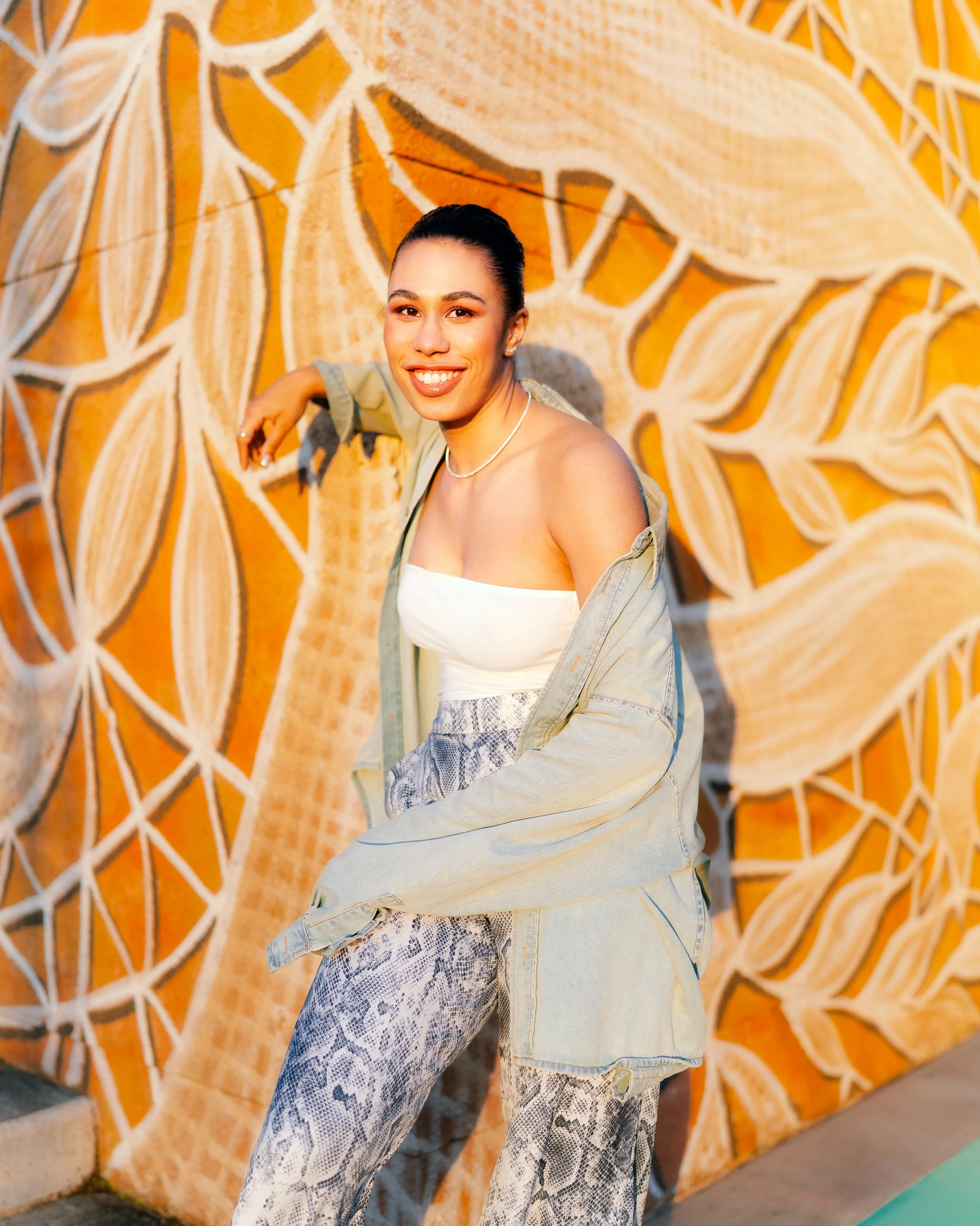 A woman smiling, wearing a white strapless top, snake-print pants, light jacket, and a pearl necklace, leaning against a colorful mural with leaf patterns.