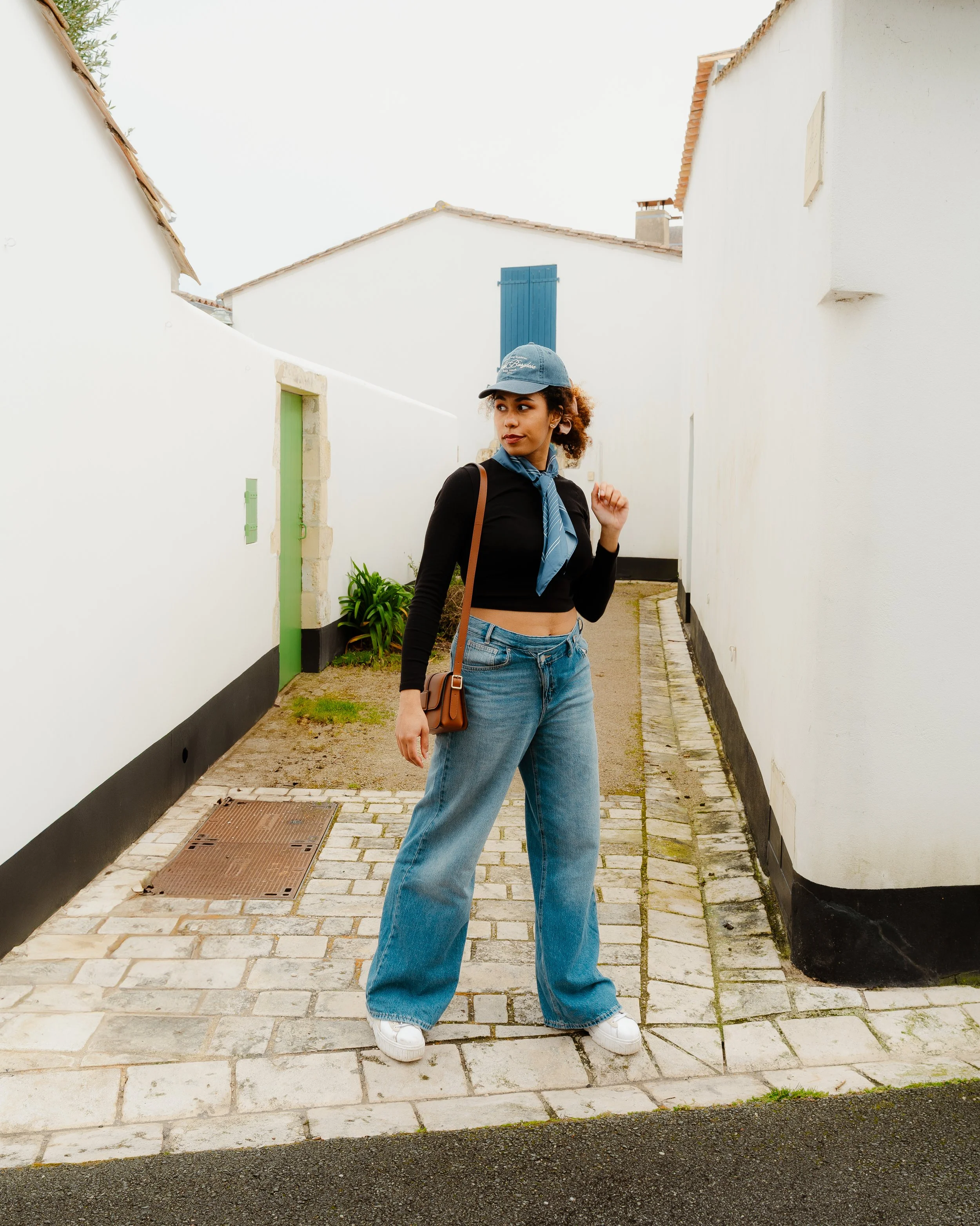 A woman with curly hair wearing a blue cap, black crop top, blue jeans, and white sneakers, standing on a small cobblestone street between white buildings with colorful doors.