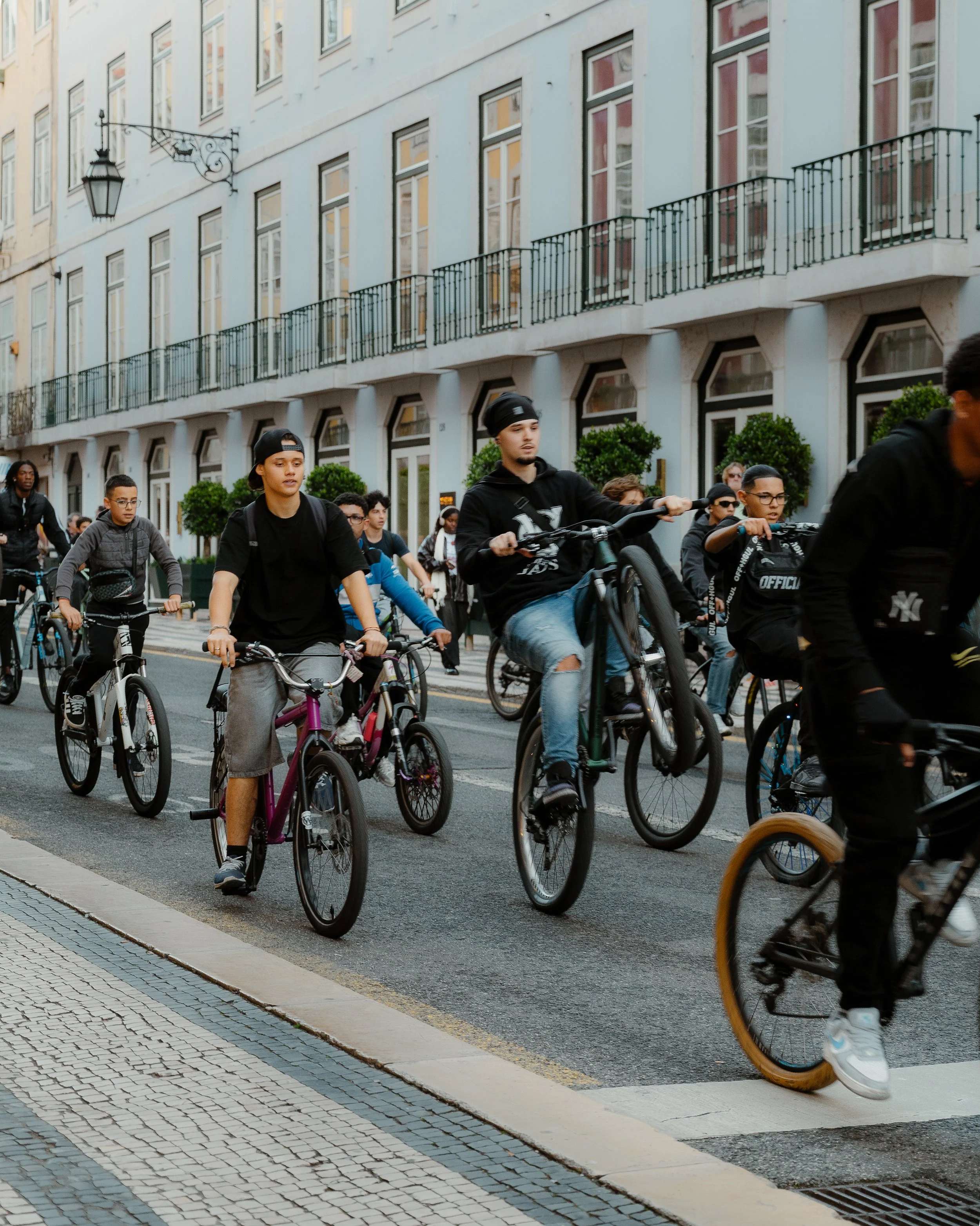 A group of people riding bicycles on a city street with a white multi-story building in the background.