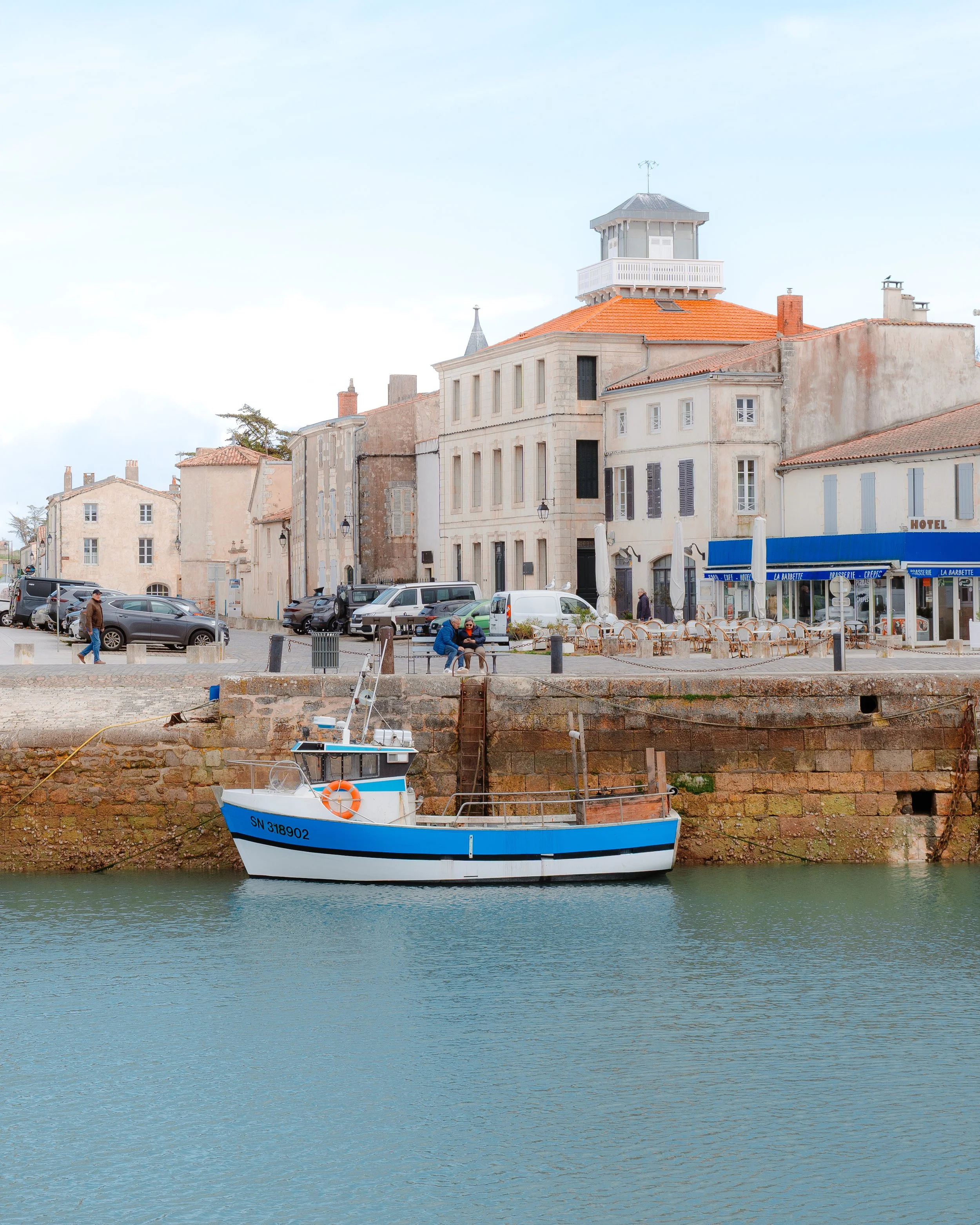 A harbor with a blue and white boat docked along a stone quay, with a town featuring beige and white buildings and parked cars in the background.