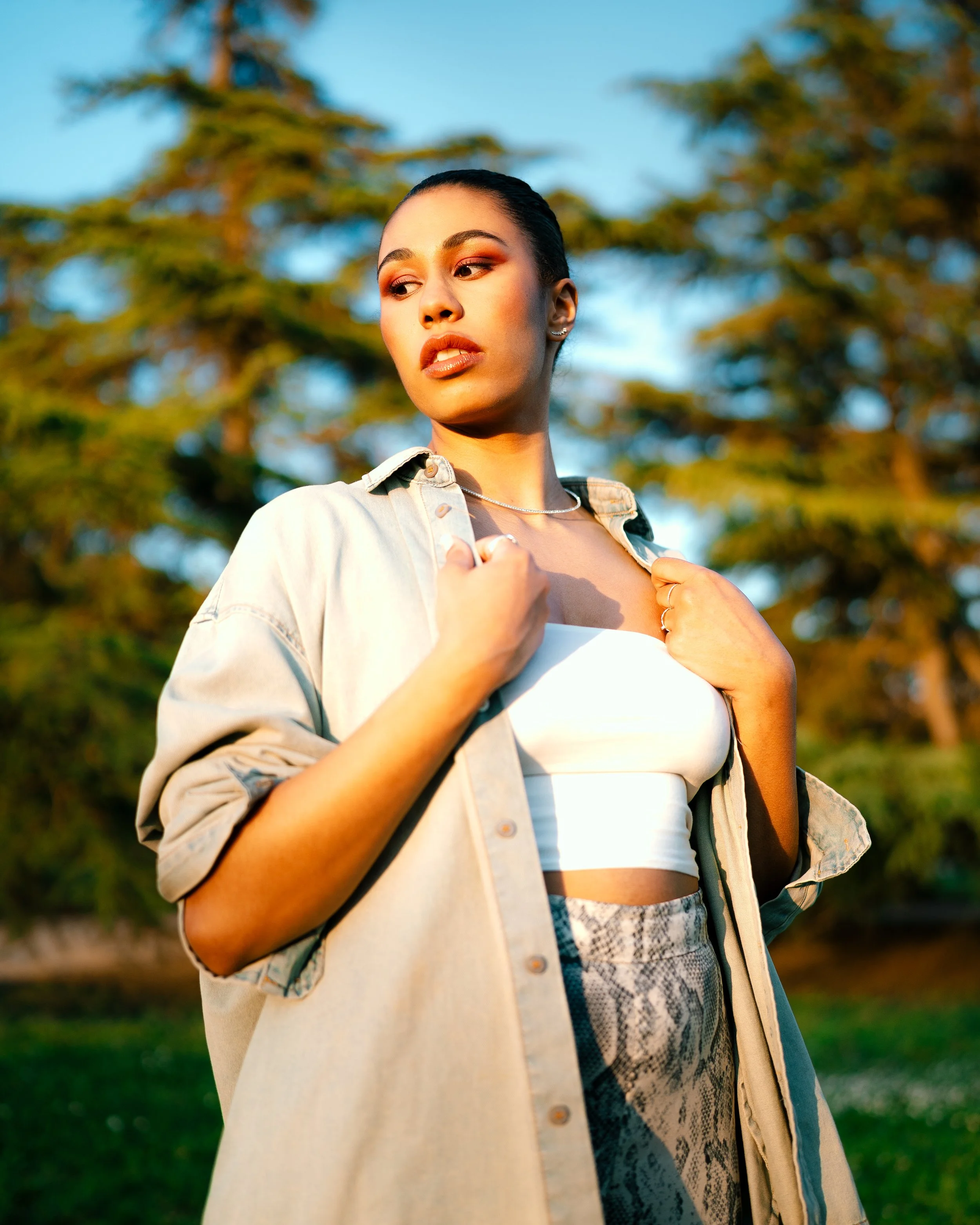 A young woman stands outdoors during golden hour, wearing a light-colored jacket over a white crop top, with her hands holding the collar. She has dark hair pulled back, minimal makeup, and is in front of trees with a blue sky.