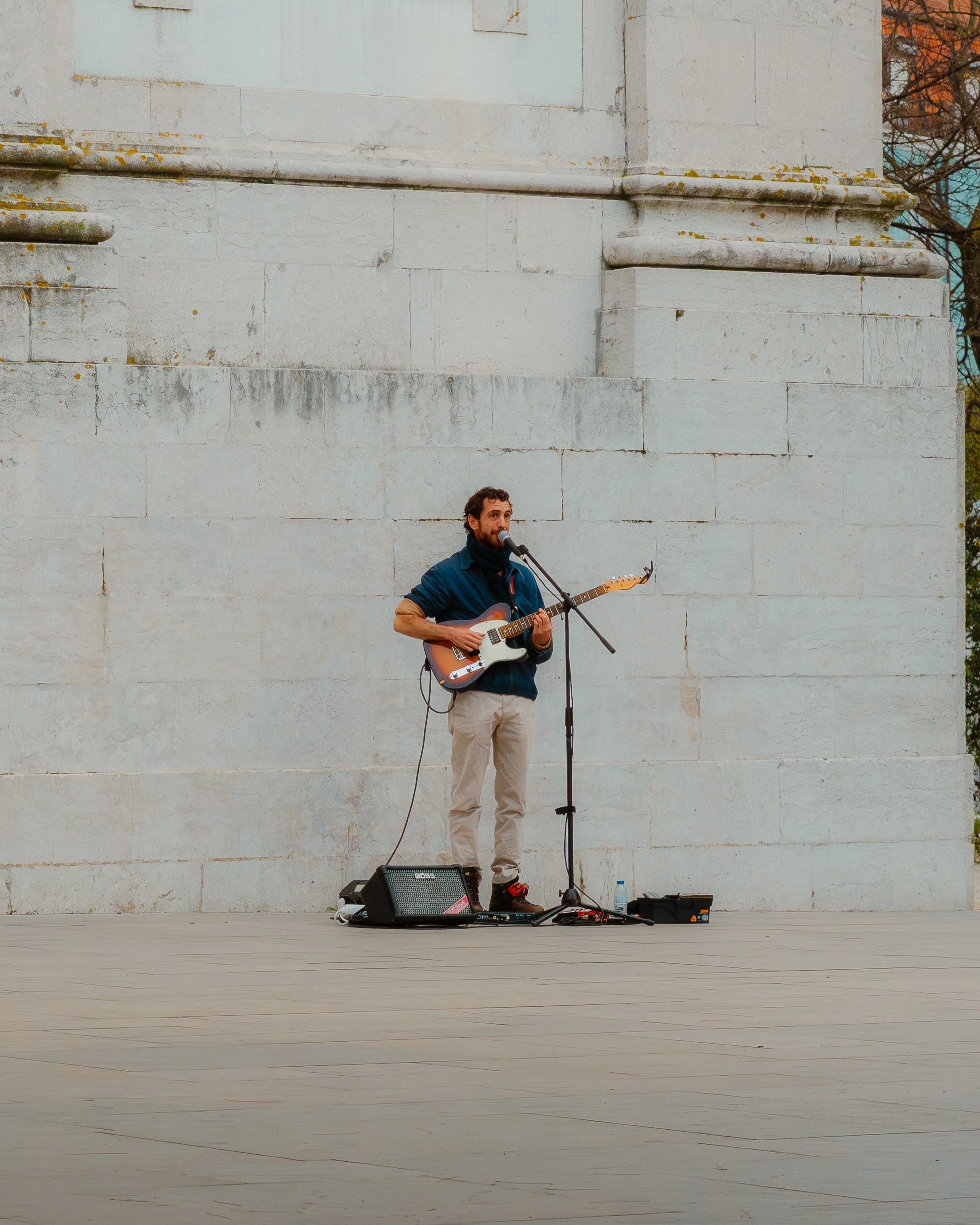A man playing an electric guitar and singing into a microphone on an outdoor stage with a large stone wall in the background.