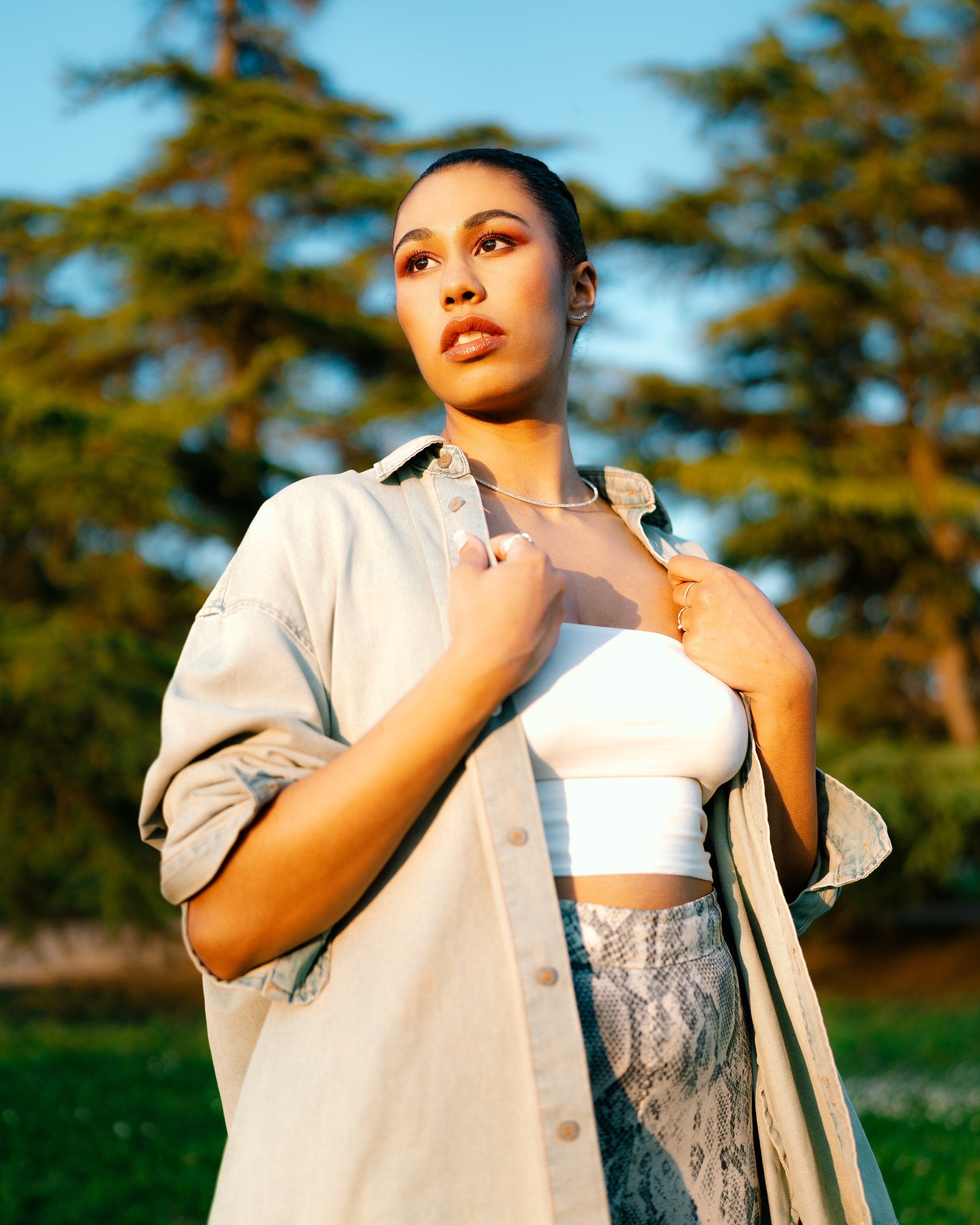 A young woman standing outdoors during golden hour, wearing a beige oversized shirt over a white crop top and patterned pants, with trees in the background.