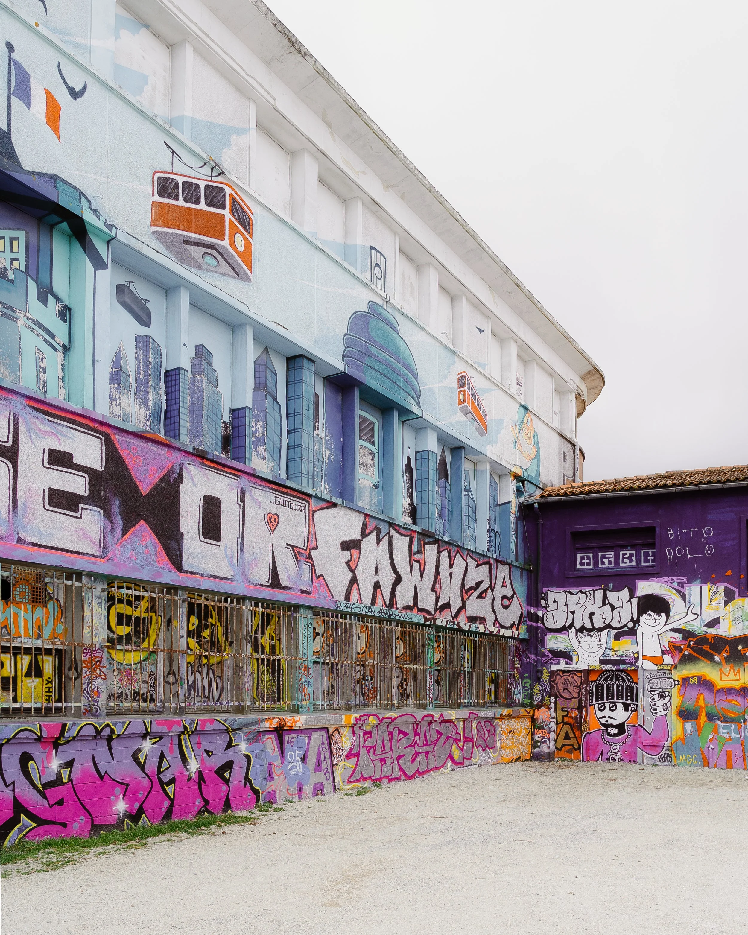 Graffiti-covered wall featuring colorful street art, cartoon characters, cityscape, and transportation motifs, with a sandy ground in front.