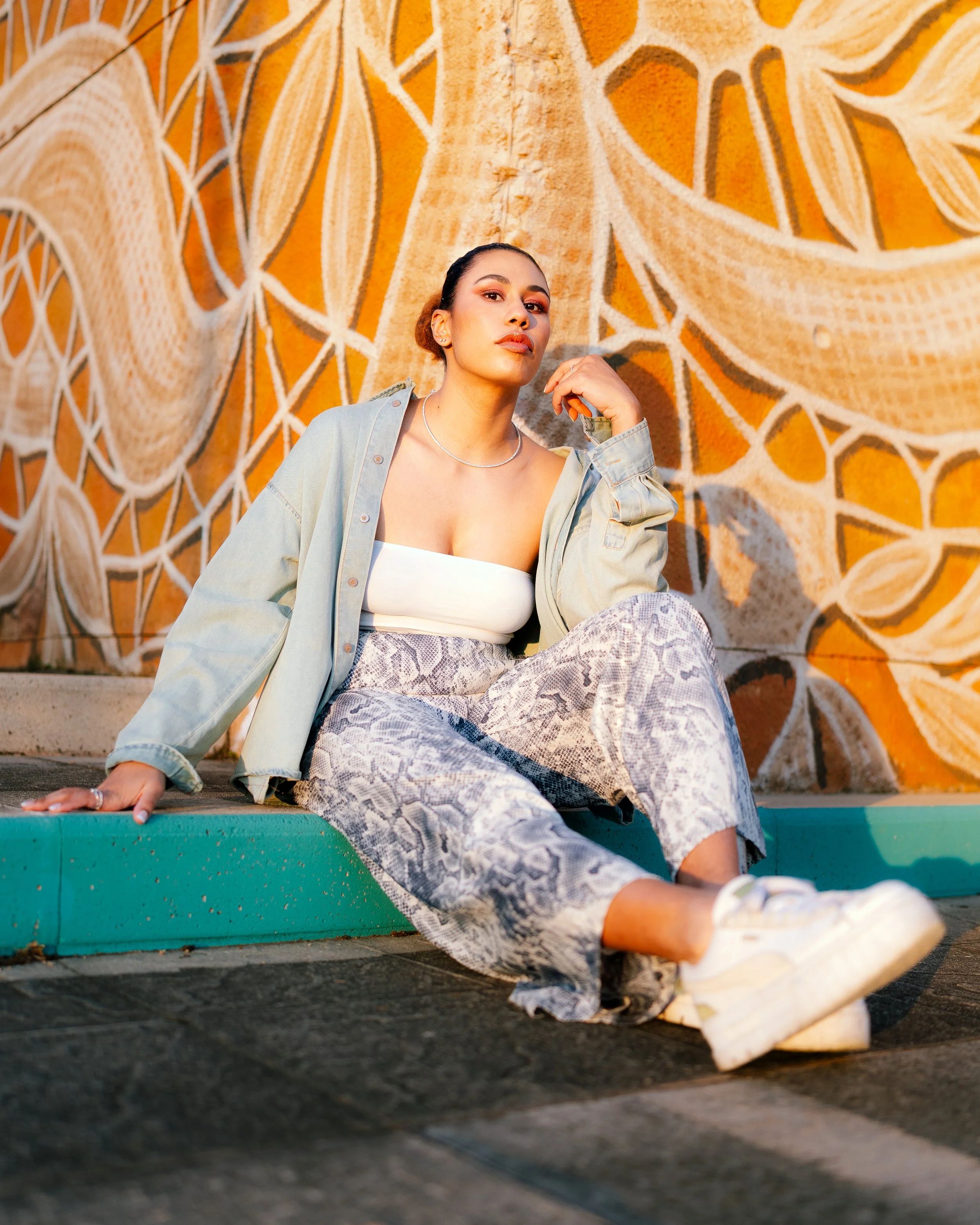 A young woman sitting on a curb in front of a colorful mural with abstract patterns, wearing a white tube top, denim jacket, snakeskin pants, and white sneakers.
