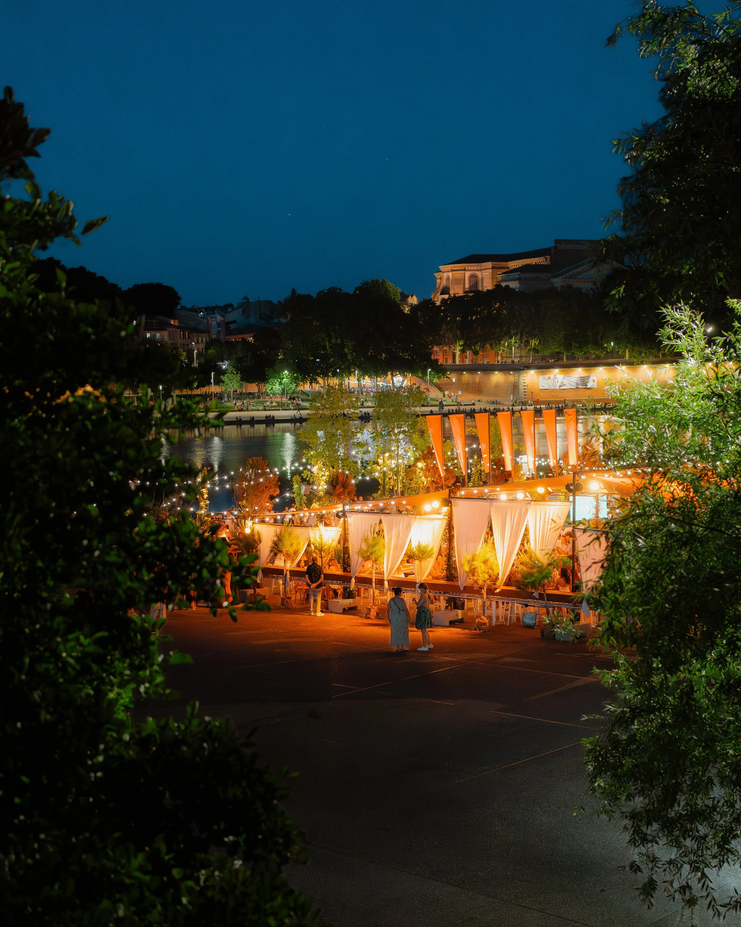 Night scene of an outdoor event with orange and white drapery, string lights, and people gathered near a waterfront with trees and city buildings in the background.