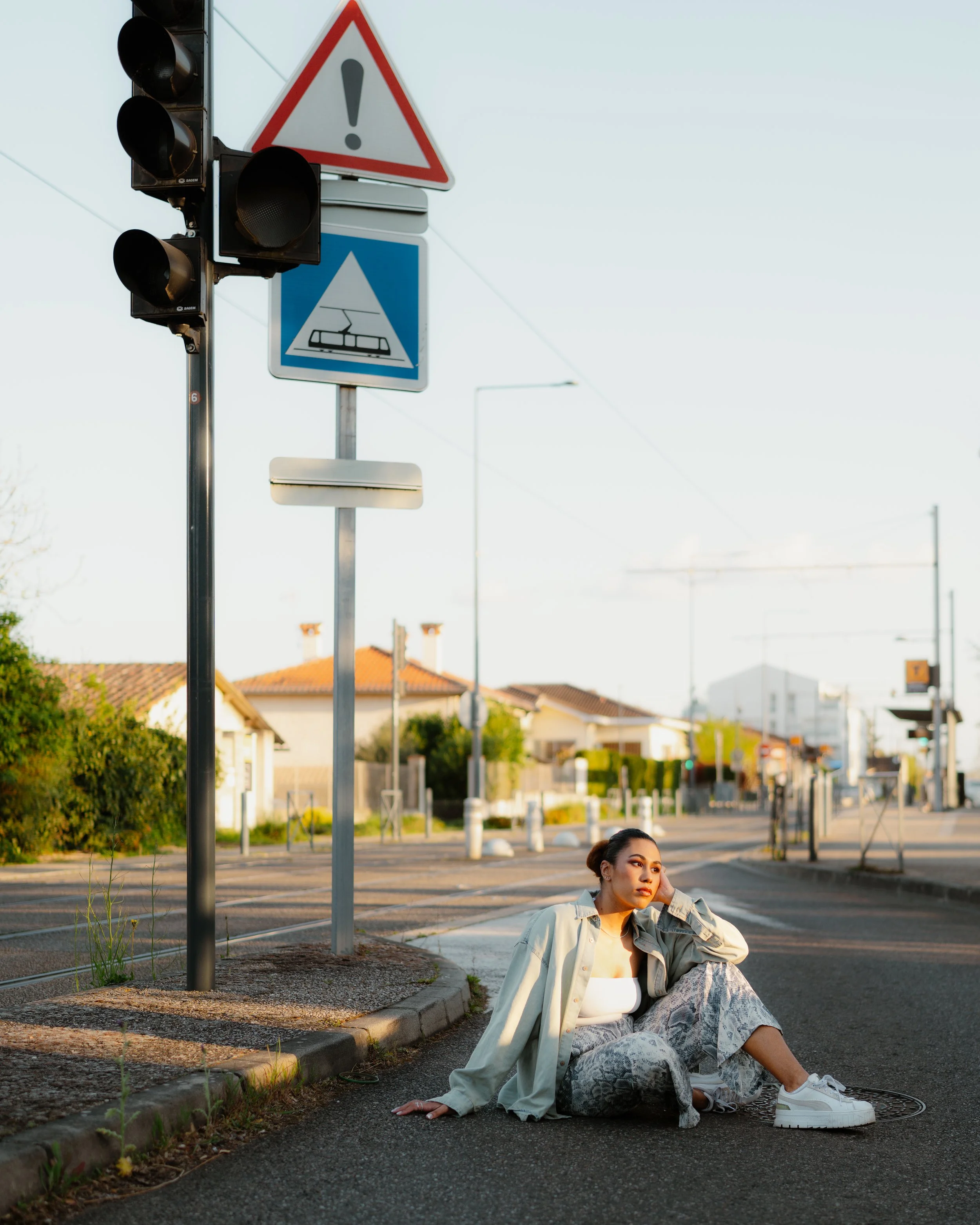 A young woman sitting on a city street near railroad tracks with traffic and warning signals, during golden hour.