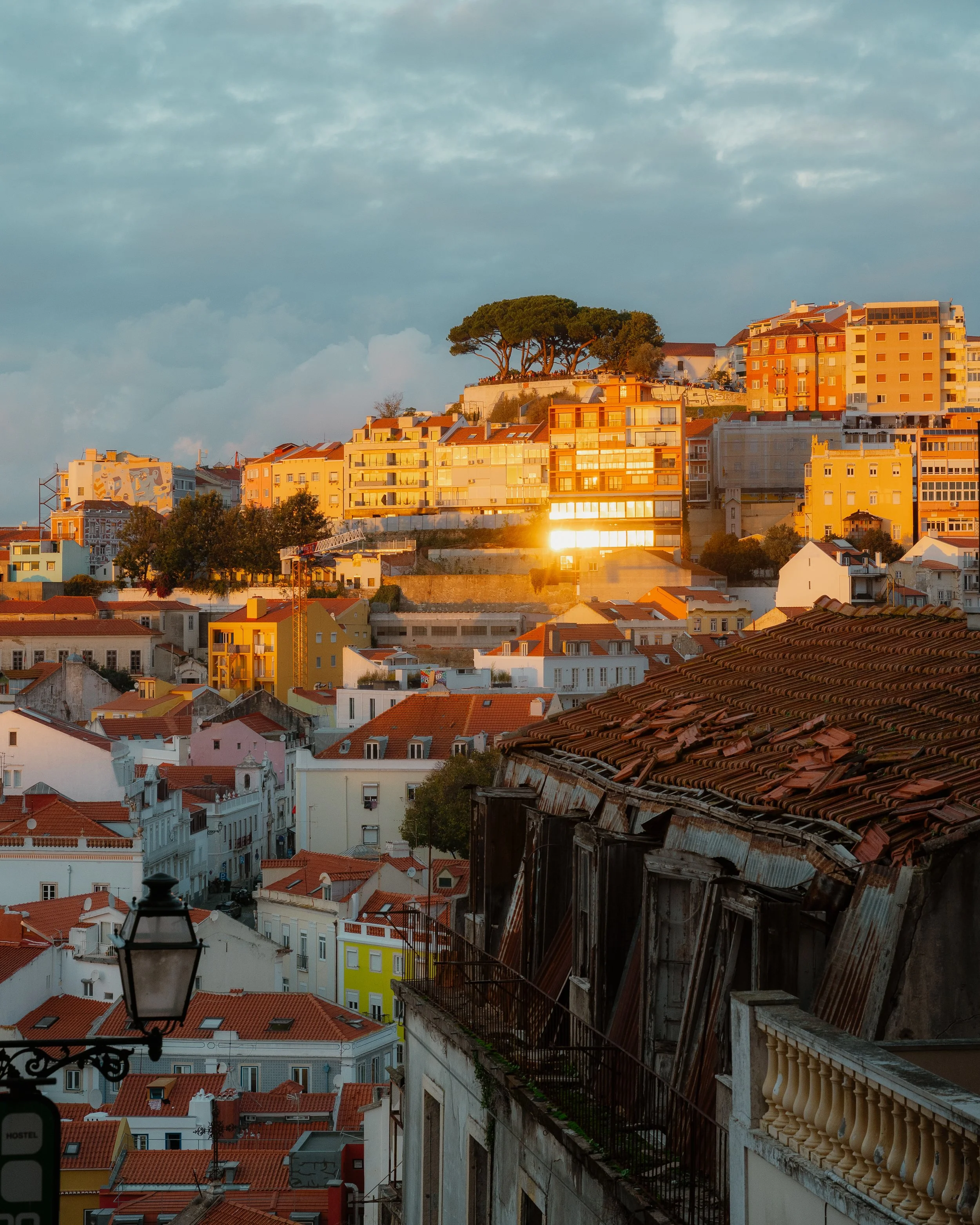 A cityscape at sunset with colorful buildings, some trees, and a cloudy sky.