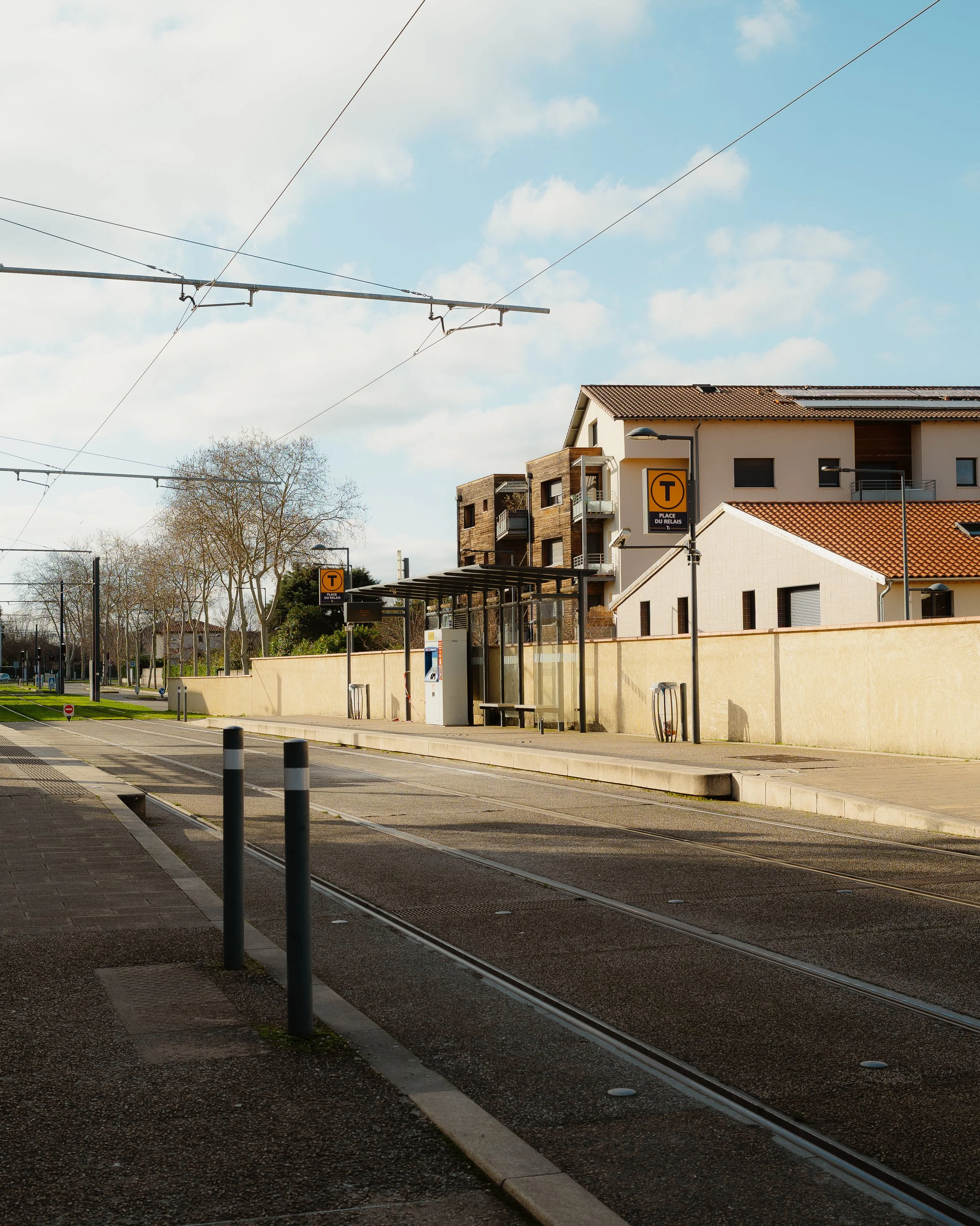 A tram stop with a shelter, ticket machine, and benches along the street, with buildings and trees in the background, under a partly cloudy sky.