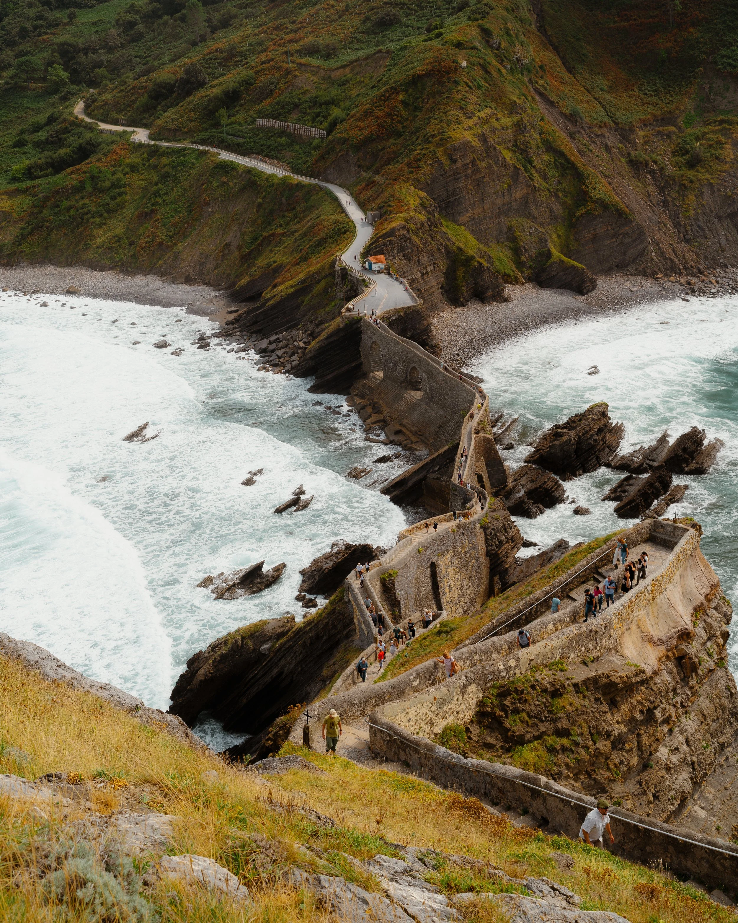 Winding coastal pathway with stairs and arches along rugged cliffs, overlooking the ocean with waves crashing against rocks.