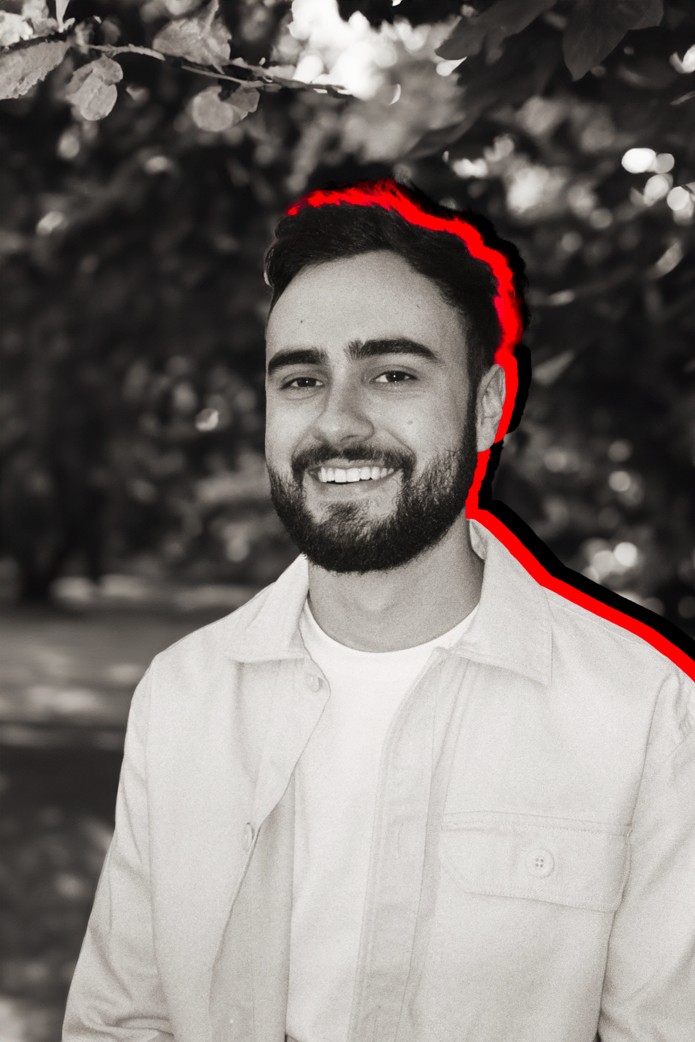 A black and white photo of a smiling man with a beard and short hair, standing outdoors with blurred trees in the background. Red outlines highlight his hair and shoulders.