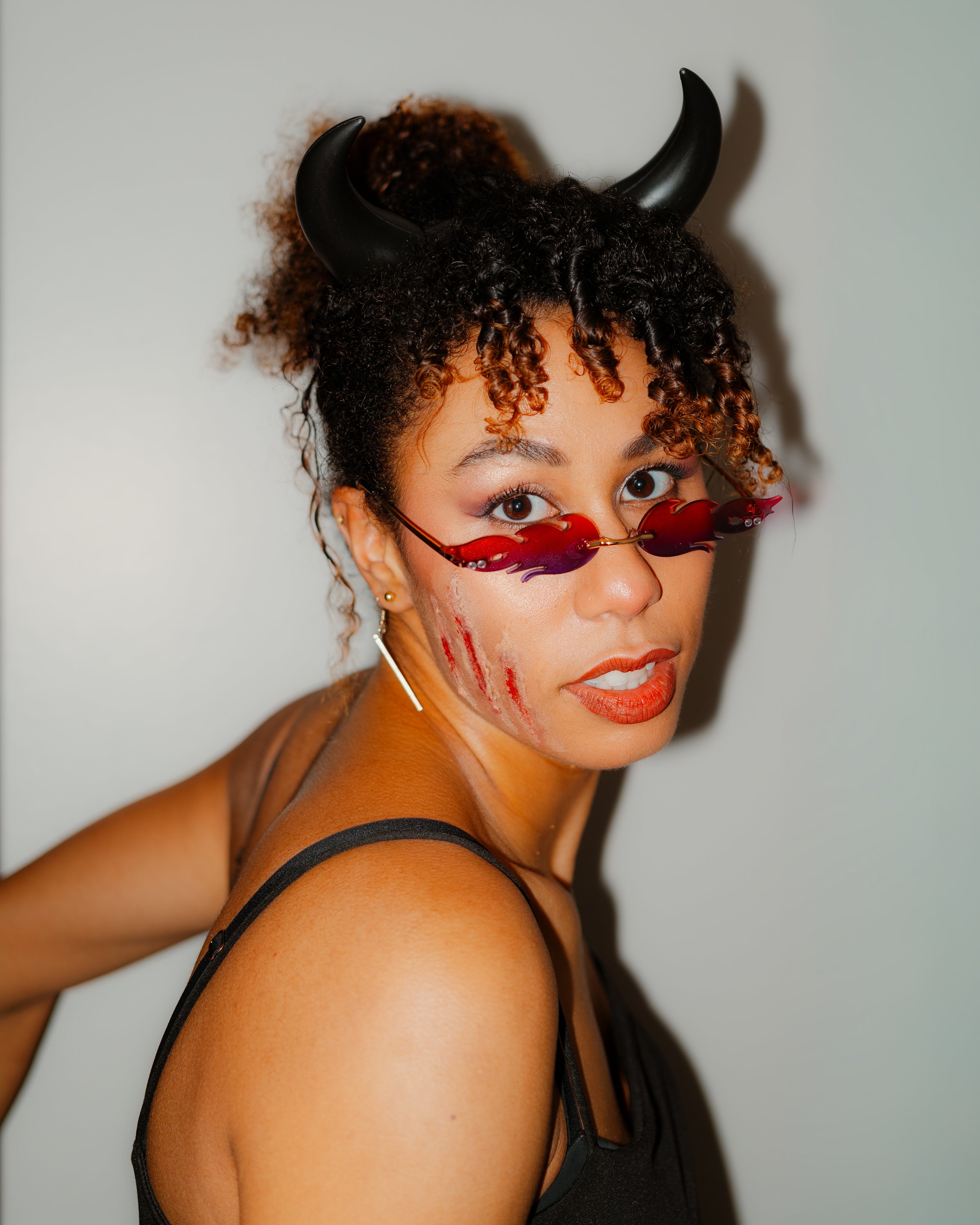 Woman with curly hair and horns wearing red sunglasses with flame design, black tank top, and faux blood scratches on face.