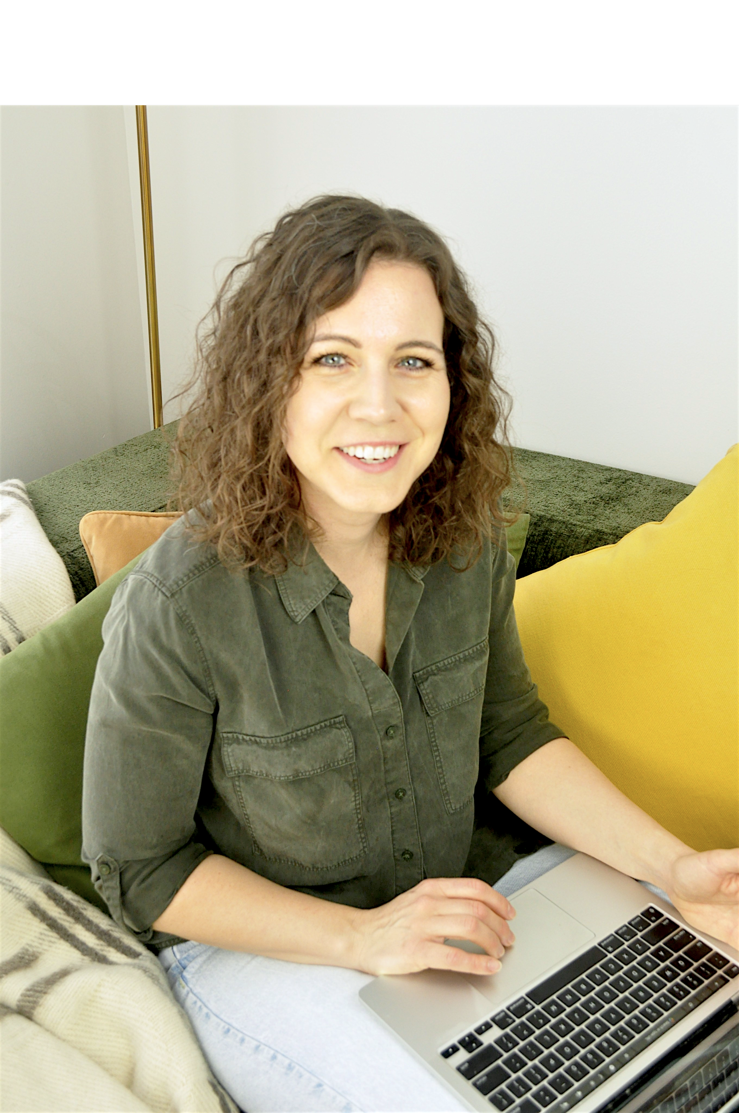 A woman with curly brown hair, light skin, and blue eyes smiling while sitting with a laptop on her lap. She is wearing a green button-up shirt and seated on a sofa with green, yellow, and beige cushions in a cozy room.