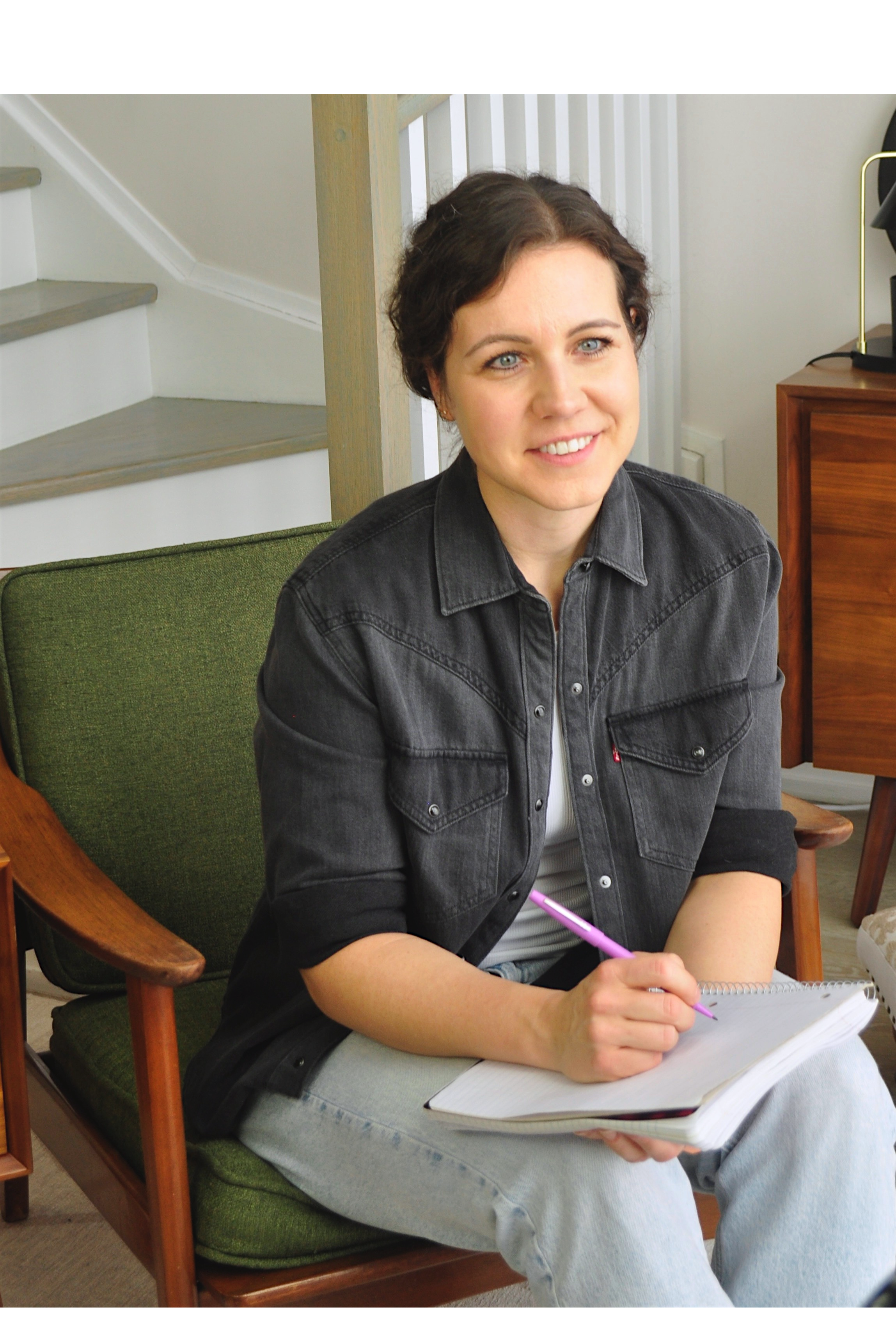 A woman with dark hair and blue eyes, wearing a black denim jacket, sitting on a green cushioned wooden chair, holding a purple pen and writing in a notebook. She is indoors with a staircase and a wooden side table visible in the background.