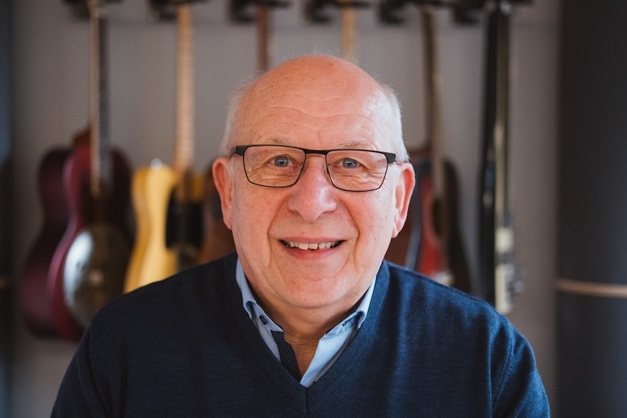 älterer Mann mit Brille und einem blauen Pullover sitzt vor einer Wand mit Gitarre in einem Musikzimmer.