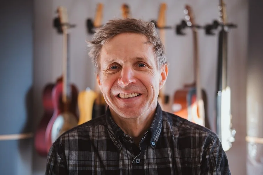 Ein Mann lächelt in einem Musikraum mit Gitarren an der Wand im Hintergrund.