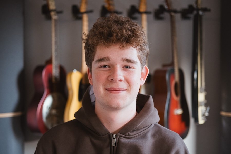 Junger Mann mit lockigem Haar vor einer Wand mit mehreren Gitarren