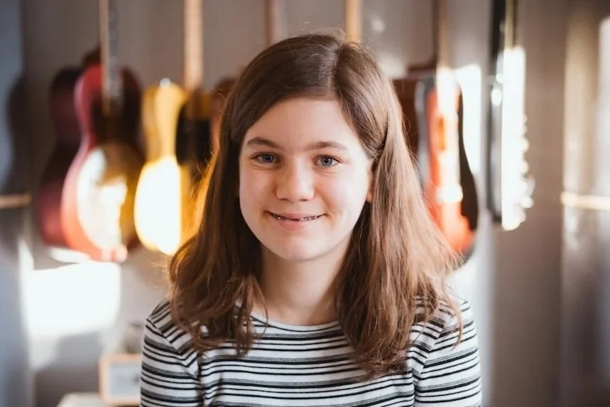 Junge Frau mit braunen Haaren und weiß-schwarzem gestreiftem Hemd, stehend vor Gitarren an der Wand, lächelnd.