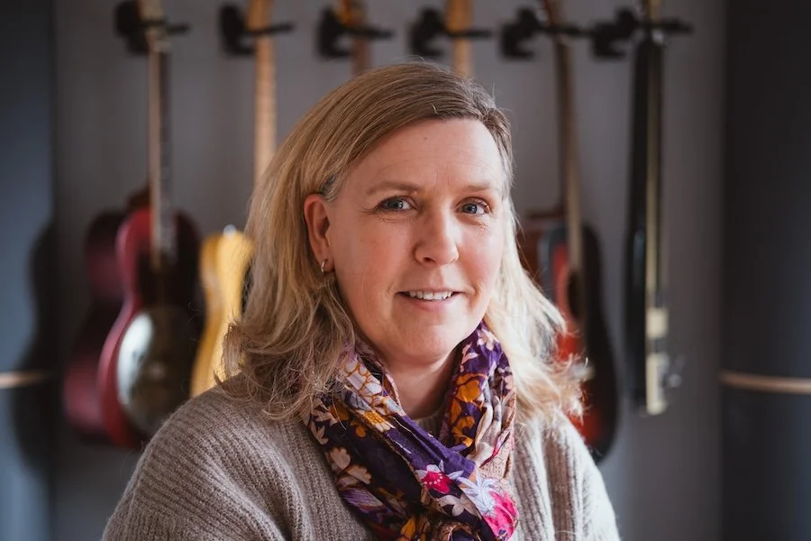 Eine Frau mit blonden, gewellten Haaren und einem bunten Schal, die vor mehreren Gitarren an einer Wand steht.