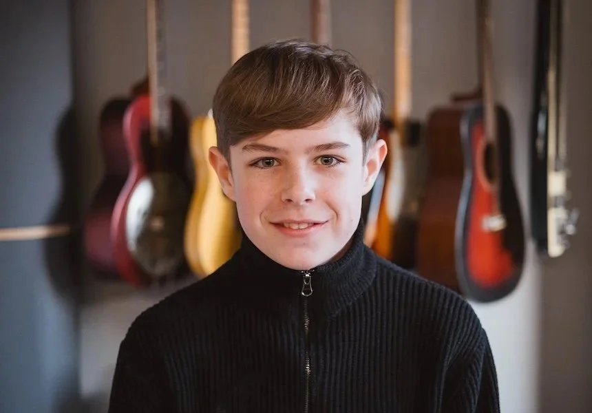 Junge Mann mit braunen Haaren in schwarzem Pullover vor einer Wand mit aufgehängten Gitarren.