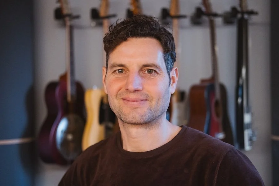 Porträt eines Mannes vor einer Wand mit mehreren Gitarren.