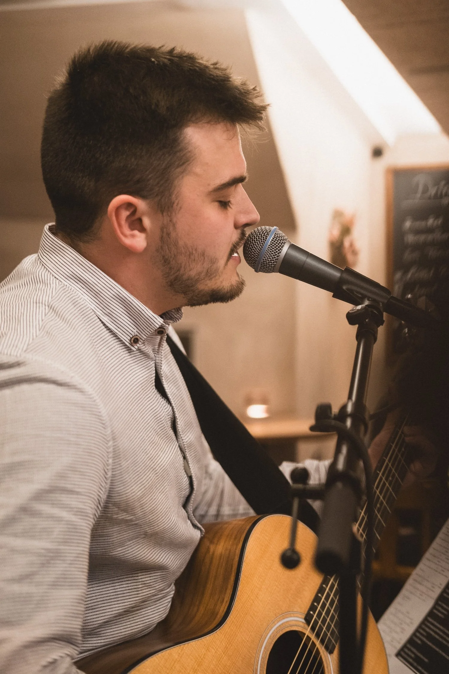 Ein junger Mann mit dunklem, kurzem Haar und Bart spielt Gitarre und singt in einem Raum, wahrscheinlich beim Live-Auftritt.