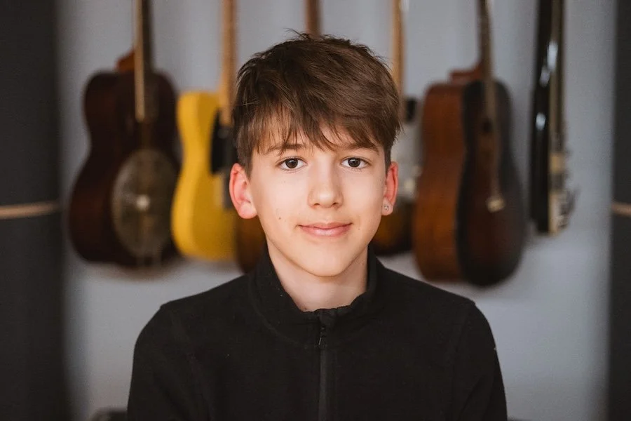 Junge Frau vor einer Wand mit mehreren Gitarren, blickt in die Kamera, trägt dunkle Kleidung, kurze braune Haare