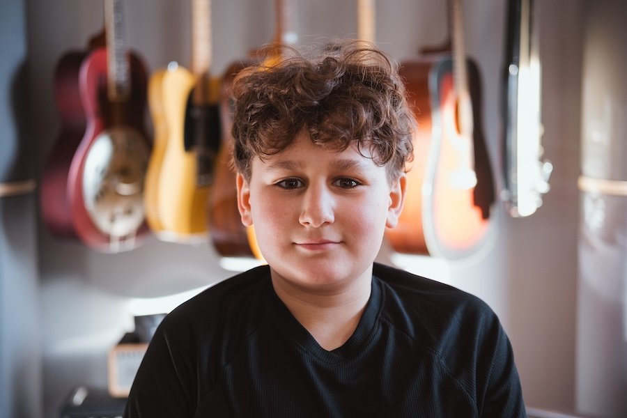 Junge mit braunen, lockigen Haaren vor einer Wand mit mehreren akustischen Gitarren.