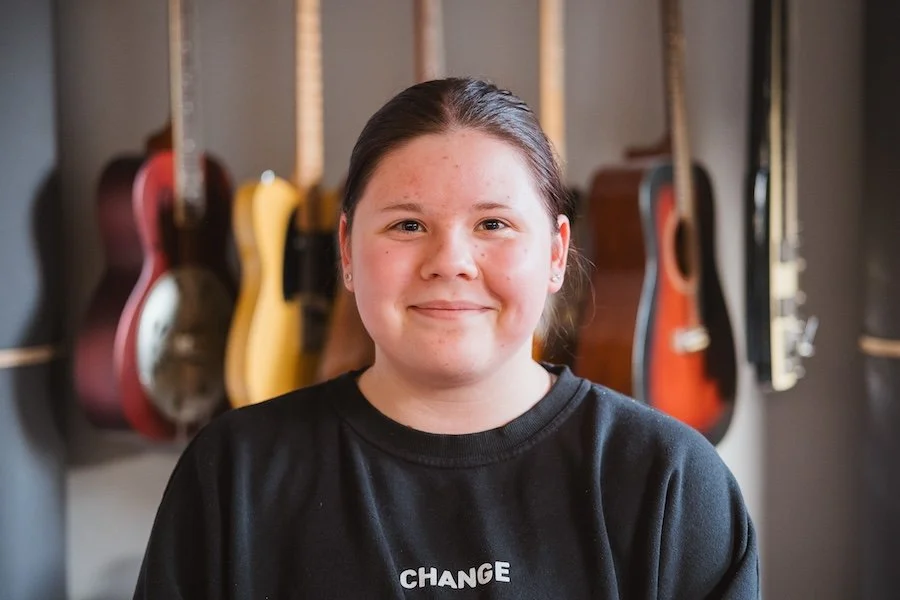 Junge Frau vor einer Wand mit Gitarren im Hintergrund, trägt ein schwarzes T-Shirt mit dem Wort 'CHANGE'.