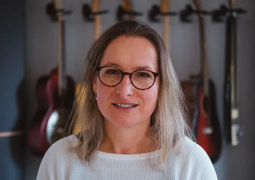 Frau mit Brille und blonden Haaren vor einer Wand mit mehreren Gitarren im Hintergrund.