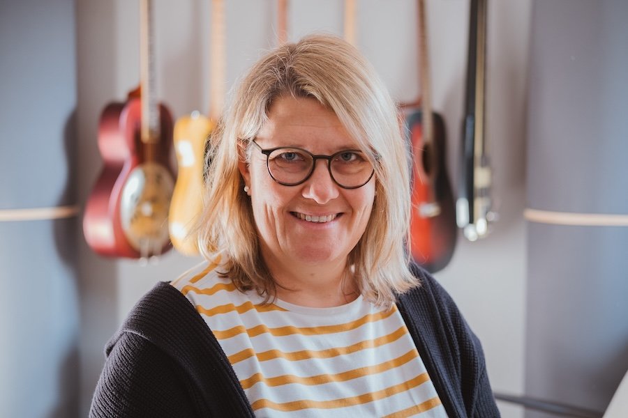 Eine lächelnde Frau mit Brille und blonden Haaren, die in einem Raum mit Gitarren im Hintergrund steht.
