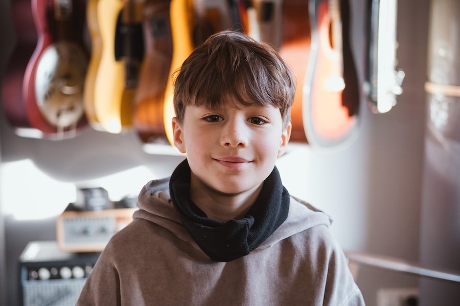 Ein Junge mit braunen Haaren steht in einem Musikgeschäft, im Hintergrund hängen Gitarren an der Wand.