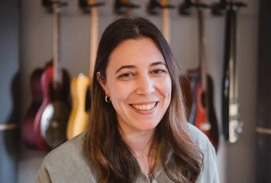 Junge Frau lächelt vor einer Wand mit aufgehängten Gitarren.