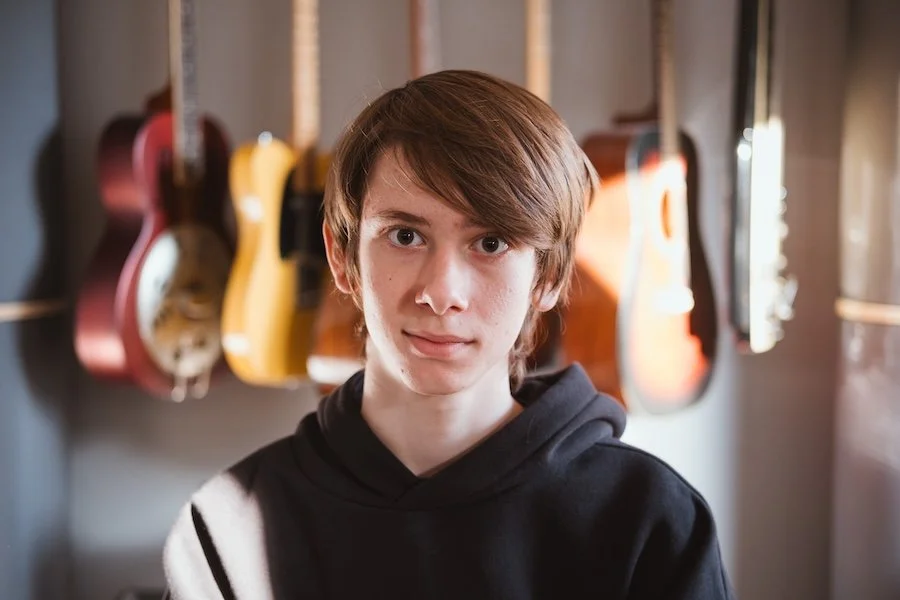 Junge mit braunen Haaren und schwarzem Hoodie vor Gitarren an der Wand.