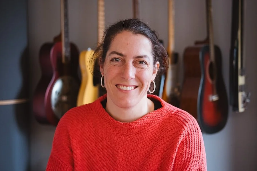 Eine lächelnde Frau mit dunklen Haaren, die eine rote Strickjacke trägt, vor einer Wand mit mehreren Gitarren.