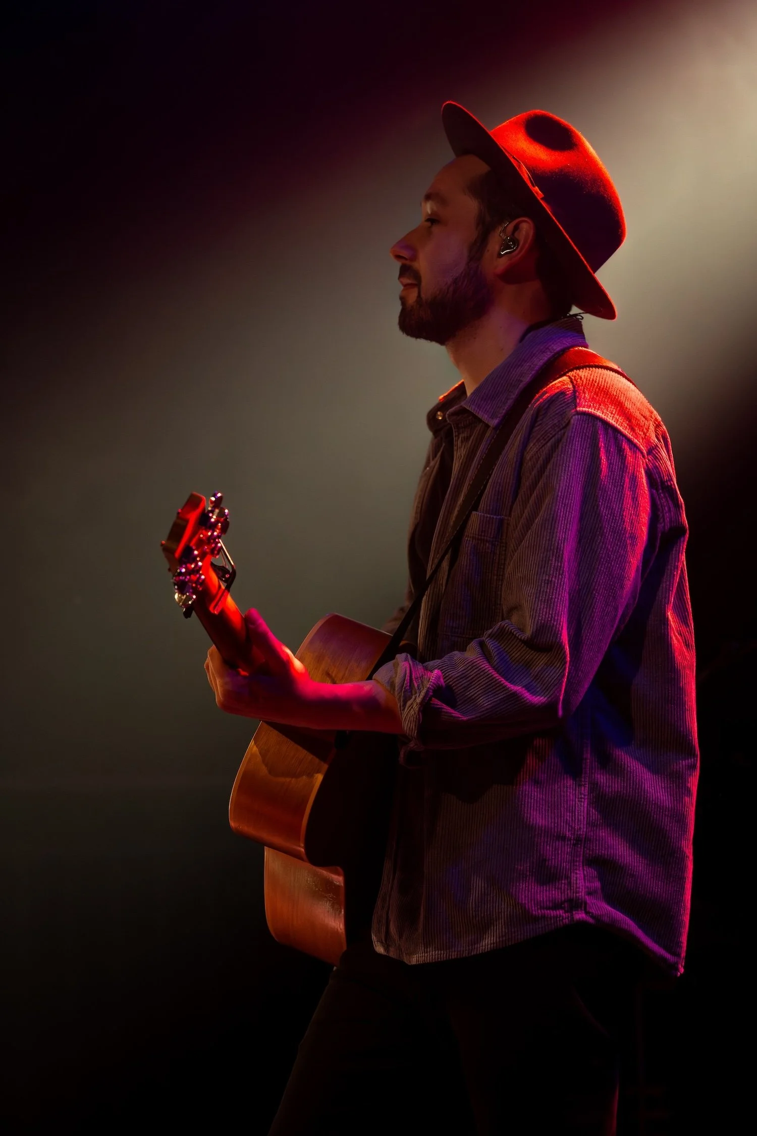 Ein Mann mit Bart trägt einen roten Hut und spielt Gitarre auf einer Bühne, beleuchtet von warmem Licht.