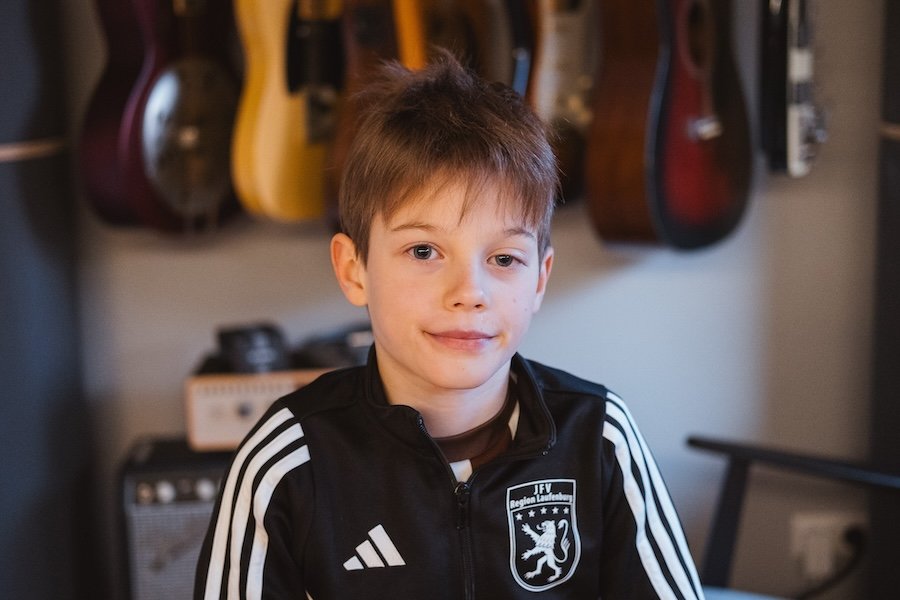 Ein Junge mit braunem Haar trägt ein schwarz-weißes adidas Trikot mit einem Vereinslogo, im Hintergrund sind Gitarren an der Wand sichtbar.