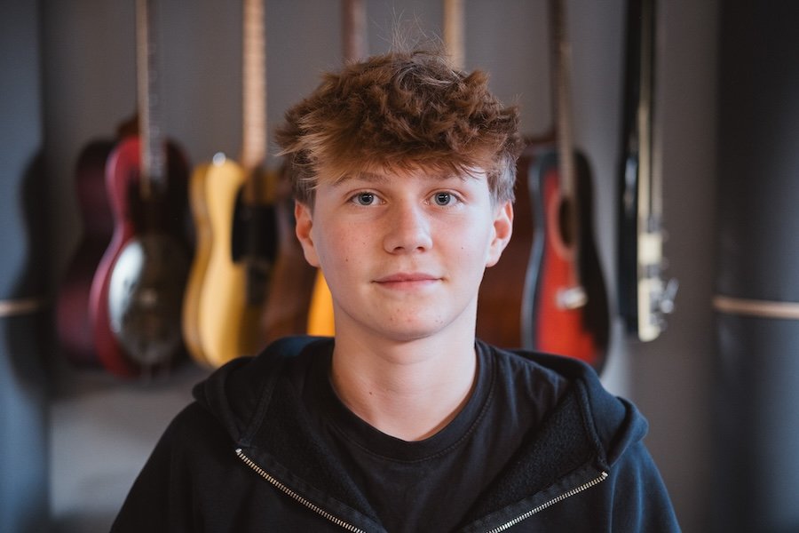 Jugendlicher Junge mit braunen, lockigen Haaren steht vor einer Wand mit mehreren akustischen Gitarren in verschiedenen Farben.