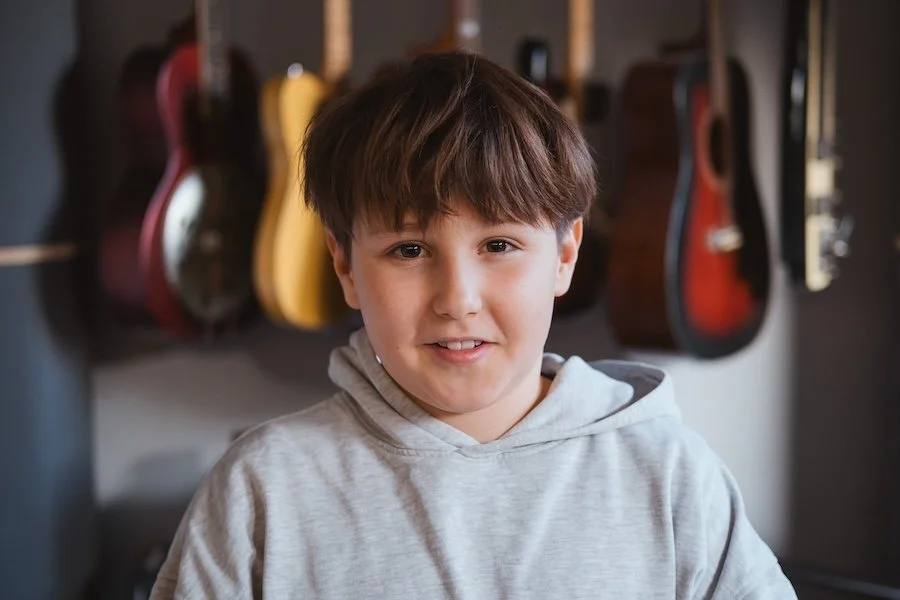 Junge mit braunen Haaren in grauem Hoodie vor einer Wand mit mehreren Gitarren.