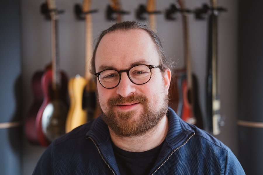 Ein Mann mit Brille und Bart vor einer Wand mit mehreren Gitarren in verschiedenen Farben.