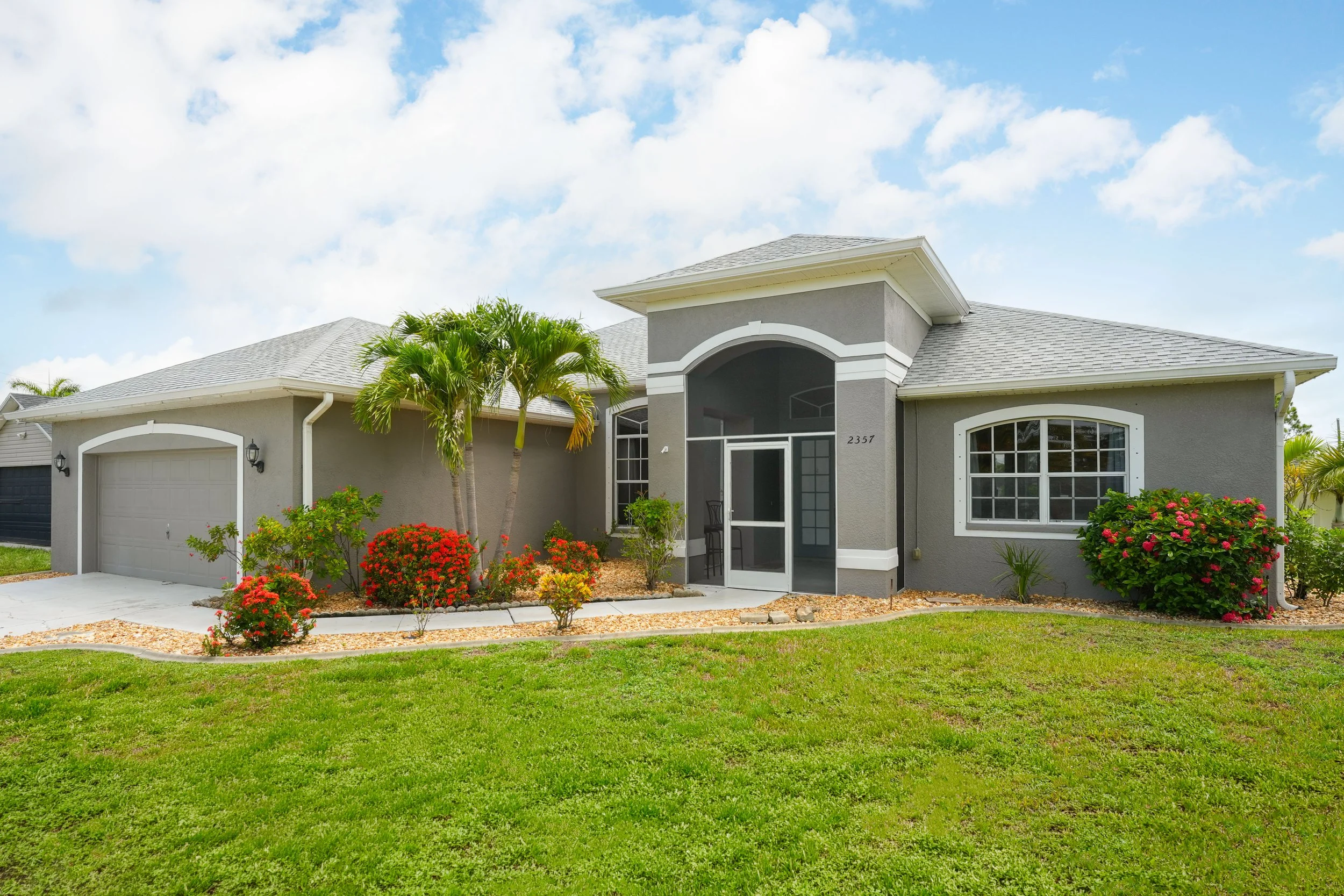 A modern gray house with white trim, a double garage, and a landscaped yard with green grass, palm trees, and colorful flowers under a partly cloudy sky.