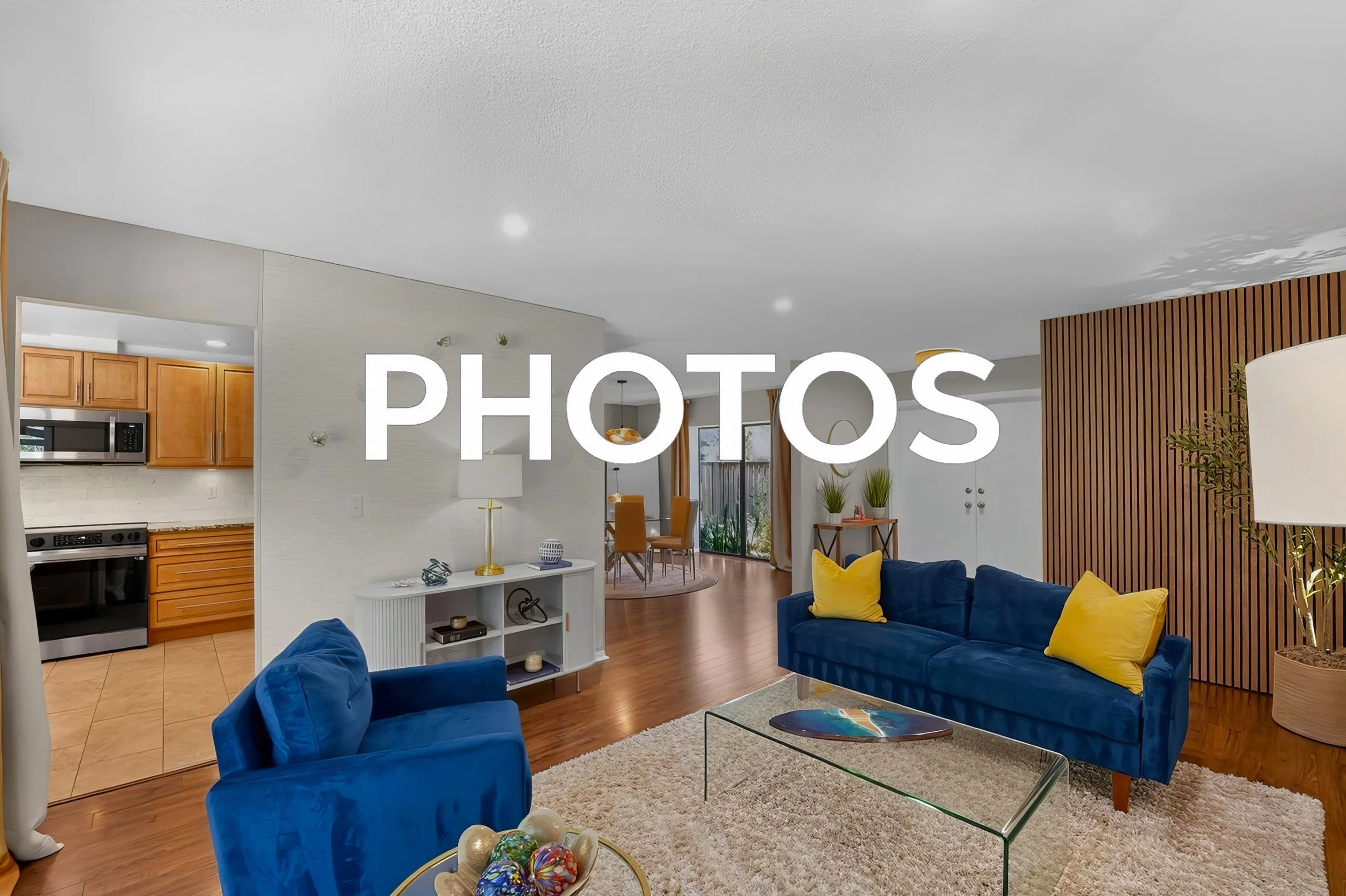 Living room with blue velvet sofas, yellow pillows, a glass coffee table, a white shag rug, a wooden dividing wall, and a view into the dining area and kitchen with wooden cabinets and stainless steel appliances.
