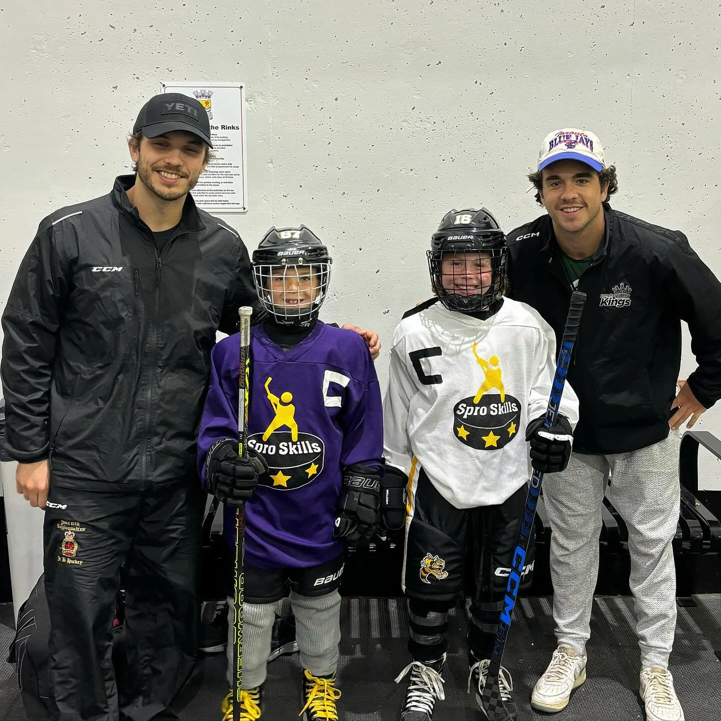 Two adult men standing next to two young boys in hockey gear, smiling indoors against a plain wall.