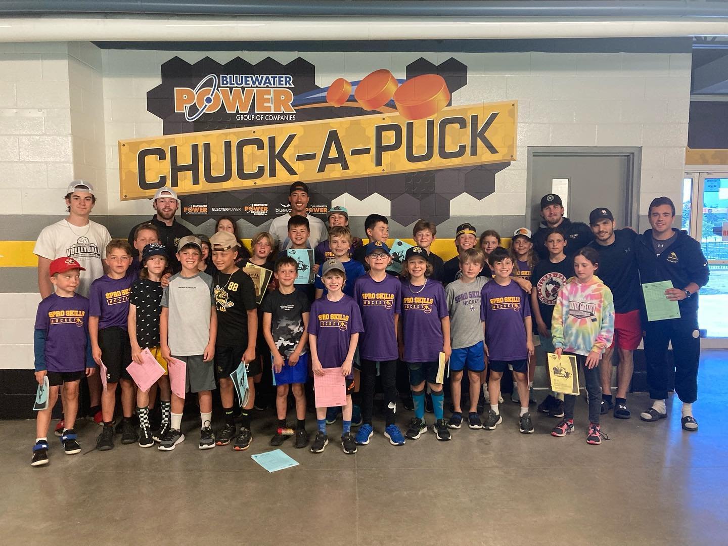 Group of children and coaches posing at an indoor hockey rink in front of a large sign that reads "Chuck-A-Puck" and "Bluewater Power" with hockey-themed graphics.