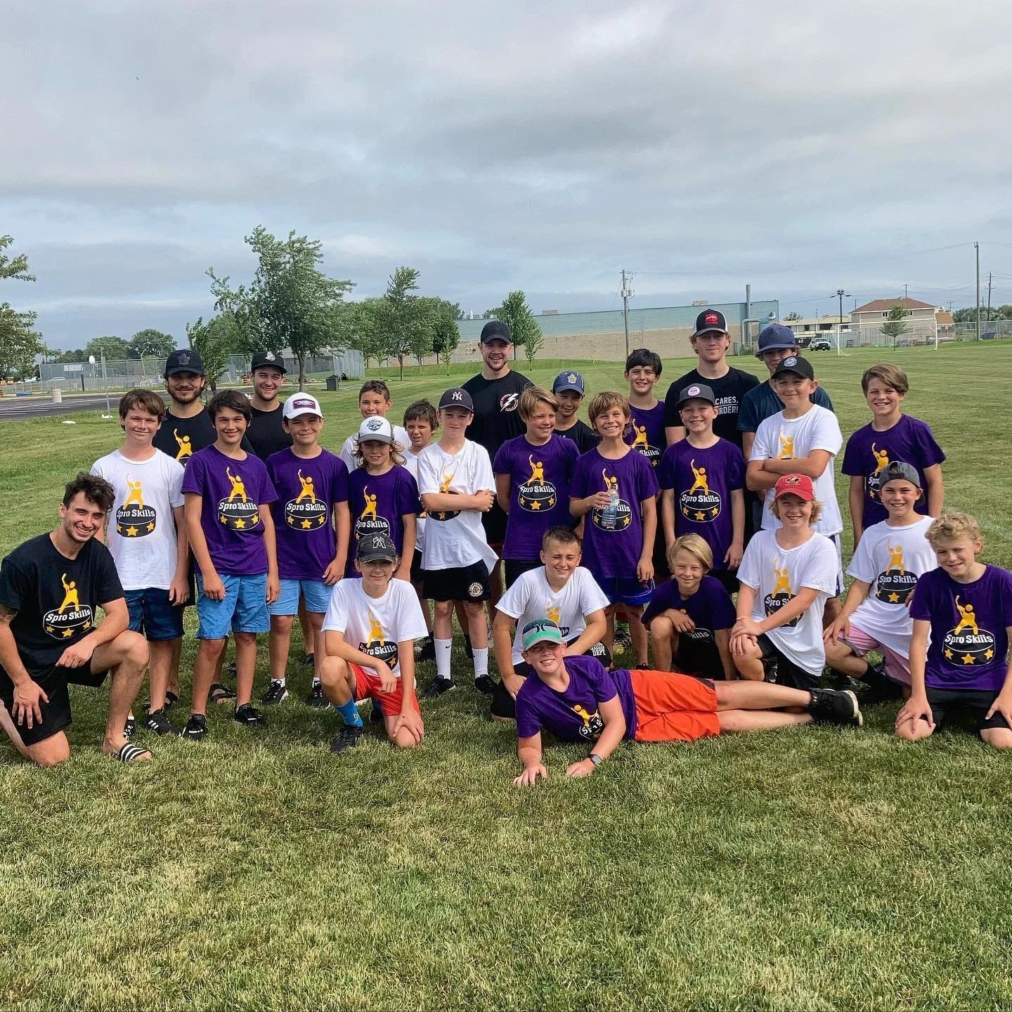 Group of children and coaches outdoors on a grassy field, some children wearing purple and white T-shirts with a logo, posing for a photo.