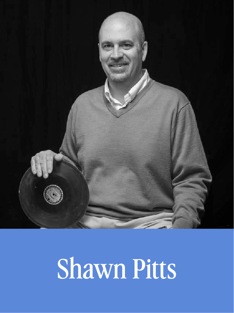 A man smiling while holding a vinyl record, sitting against a dark background, with a blue banner that says "Shawn Pitts".