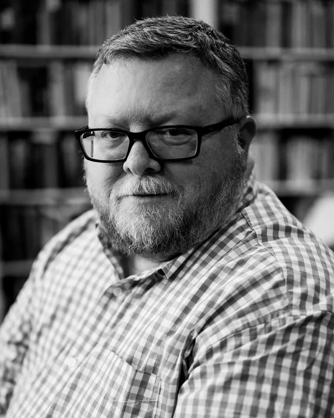 Black and white portrait of a man with glasses and a beard, wearing a checkered shirt, in front of a bookshelf.
