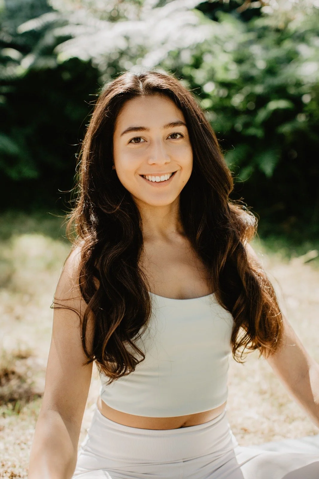 A young woman with long wavy dark hair smiling outdoors in sunlight, wearing a white sleeveless crop top and matching pants, with greenery in the background.