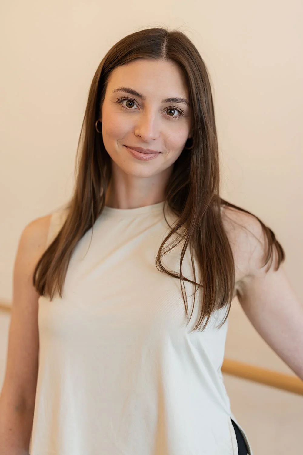 A woman with long brown hair, wearing a cream sleeveless top, smiling in front of a plain light-colored wall.